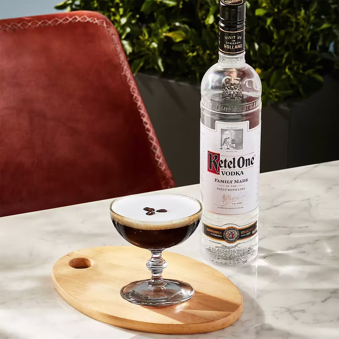 A bottle of Ketel One vodka stands next to an espresso martini garnished with coffee beans on a wooden board atop a marble table.