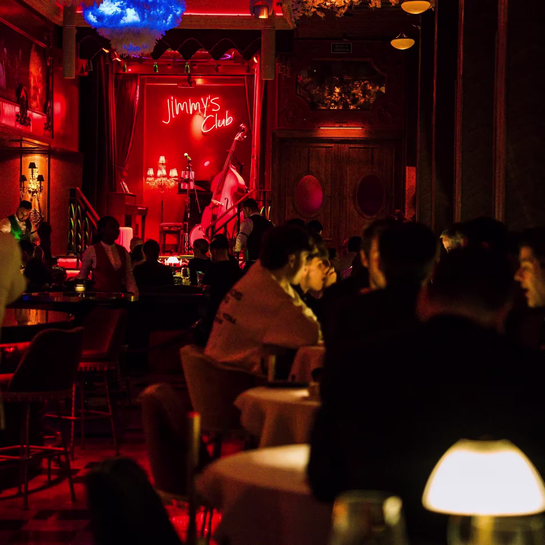 Jimmy´s: El interior de Jimmy´s, en el  fondo de la sala se observa un violonchelo en un escenario pequeño, acompañado de un letrero con luces de neón, hay multiples mesas ocupadas por personas y muchos elementos florales y lámparas decorativas.