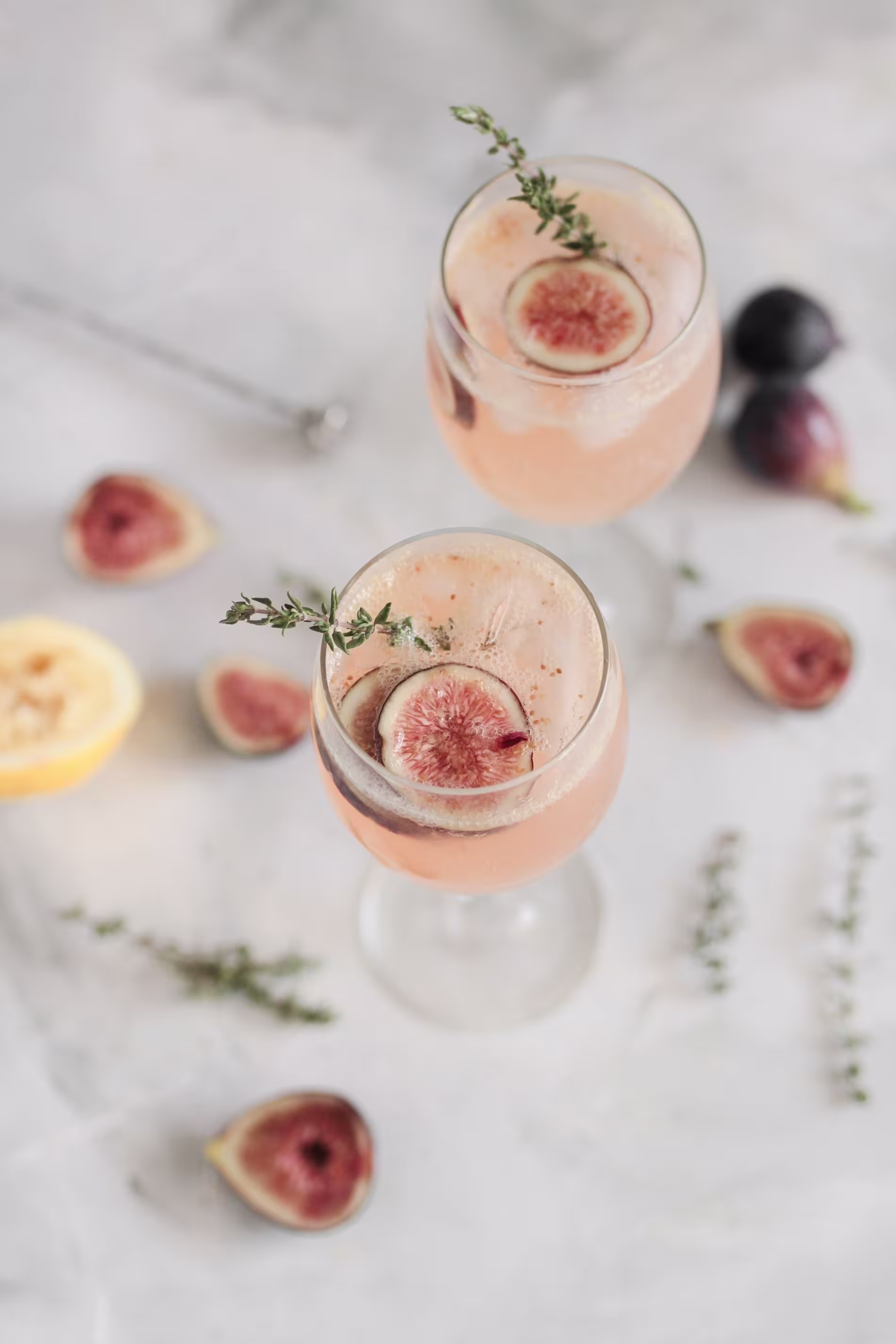 Two stemmed glasses of a light pink beverage are garnished with fresh fig slices and sprigs of thyme, surrounded by additional figs and herbs on a light surface.