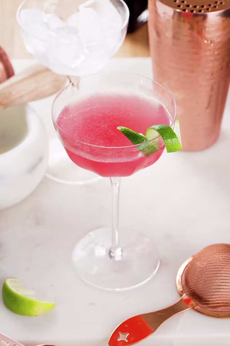 La imagen presenta una copa con el cóctel Cosmopolitan y otros elementos de coctelería.