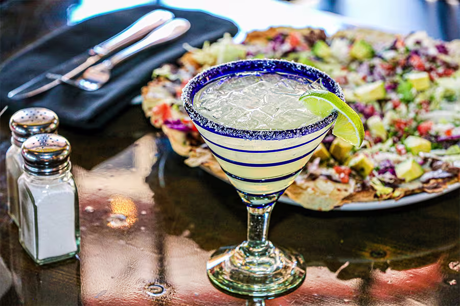 A margarita with a lime wedge sits on a table next to a plate of nachos, salt and pepper shakers, and a set of utensils wrapped in a napkin.