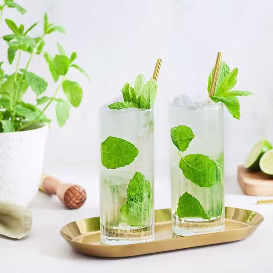 En la foto se ven dos vasos con el cóctel Mojito, y en el fondo, hay elementos de coctelería relacionados con este trago.