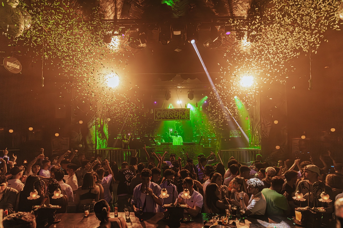 La imagen muestra una fiesta animada en un club nocturno con una multitud de personas disfrutando frente a un DJ. Confeti cae del techo mientras luces verdes y doradas crean un ambiente festivo y lleno de energía.