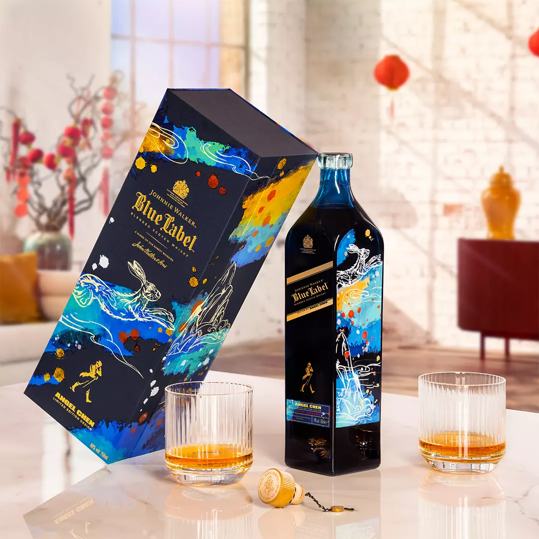 A limited edition Johnnie Walker Blue Label bottle with a decorated box, two whiskey glasses with liquor, and a cork on a table in a bright, modern room.
