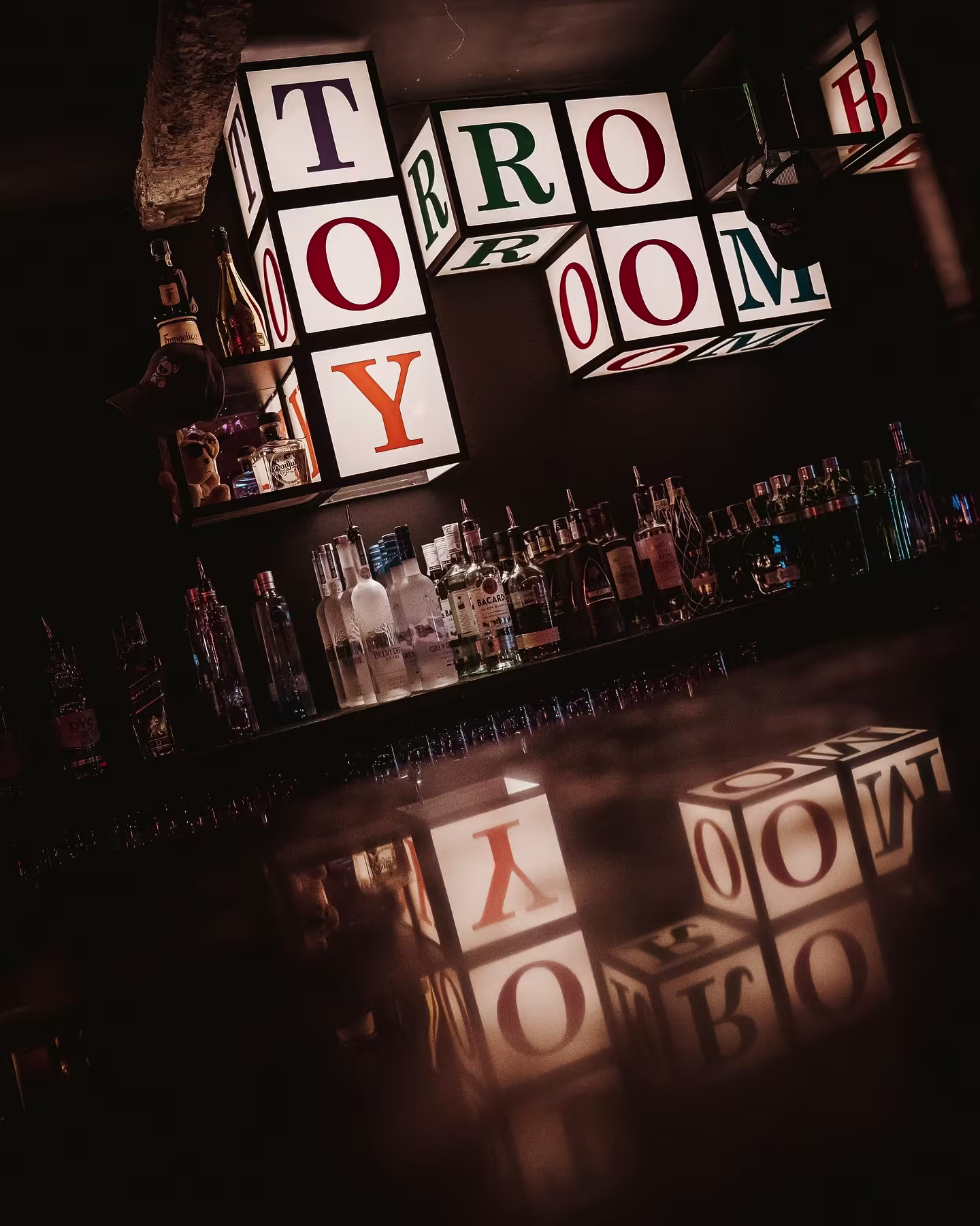 La imagen de TOY ROOM muestra un ambiente vibrante con luces de colores y una barra llena de botellas, creando una atmósfera energética y moderna.