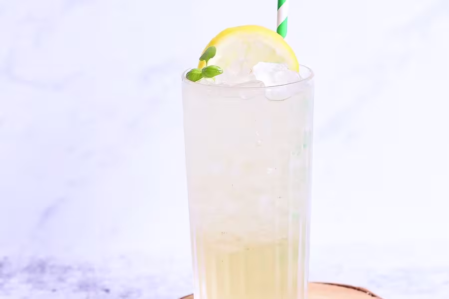 A tall glass of iced lemonade garnished with a lemon slice and green sprig, topped with a green-striped straw, on a light surface.