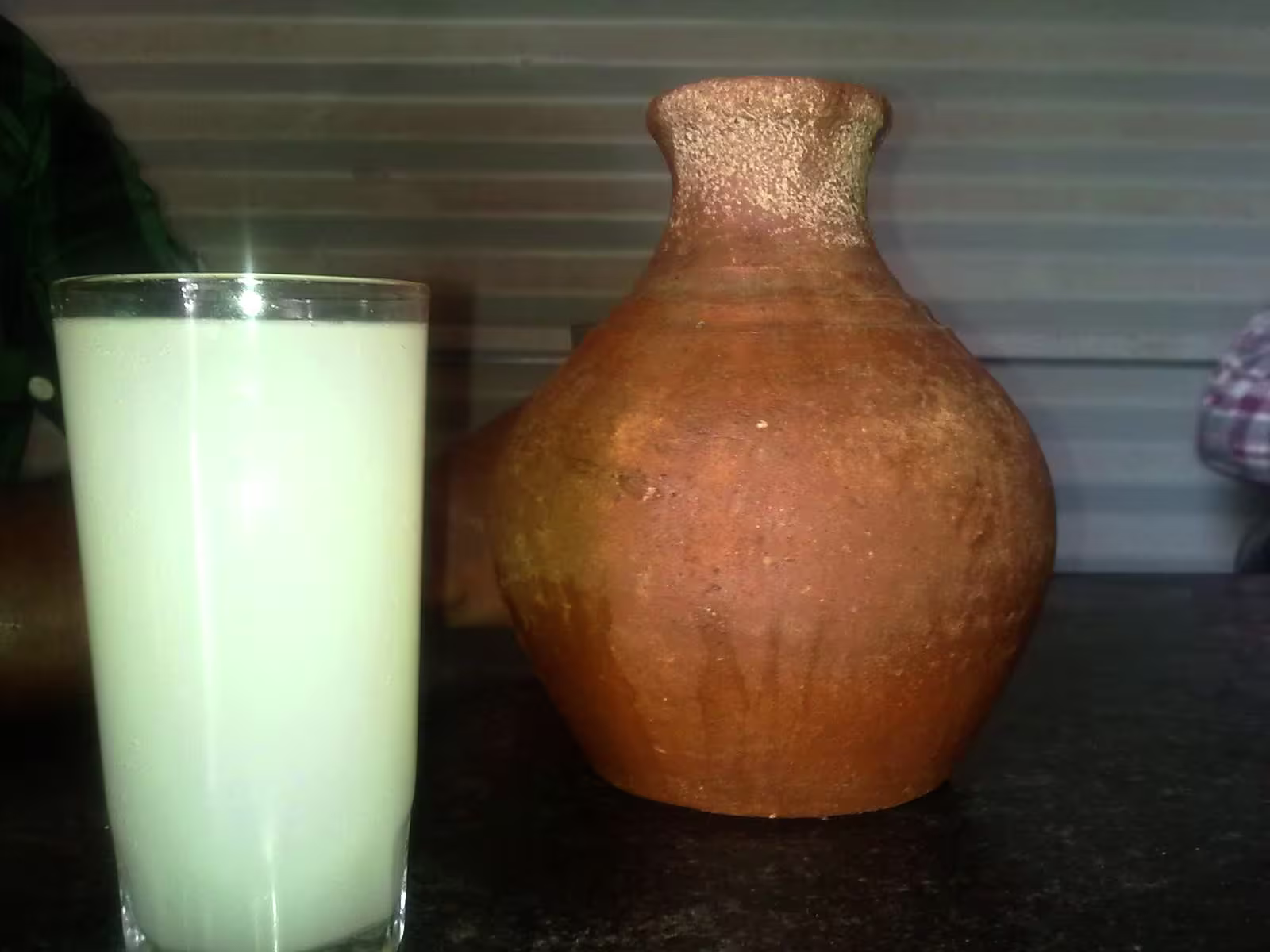A glass of white, opaque liquid sits next to a brown, round clay pot on a dark surface with a striped background.