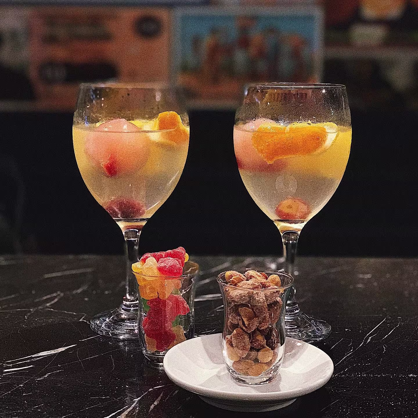 Livingstone: Dos copas frescas con frutas cítricas acompañadas de dulces y frutos secos, dispuestas sobre una mesa oscura. Un ambiente acogedor para disfrutar de bebidas con un toque frutal y aperitivos.