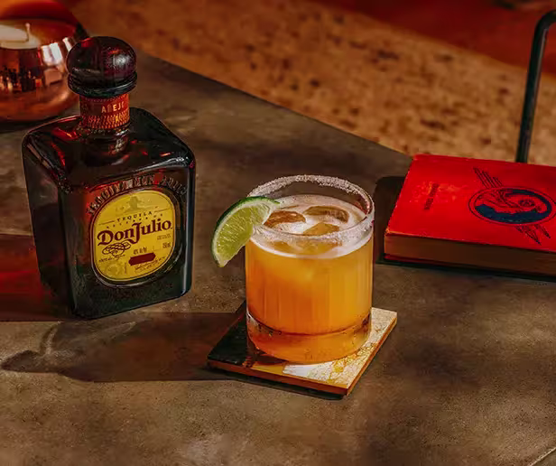 A glass of cocktail with a lime wedge sits on a coaster next to a bottle of Don Julio tequila and a closed red book on a table.