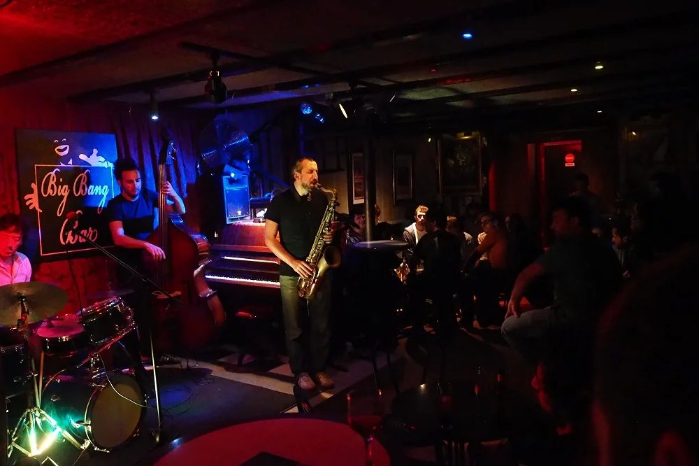 Concierto de jazz en vivo en Big Bang Bar, con músicos tocando batería, contrabajo y saxofón, rodeados de un público atento en ambiente íntimo.