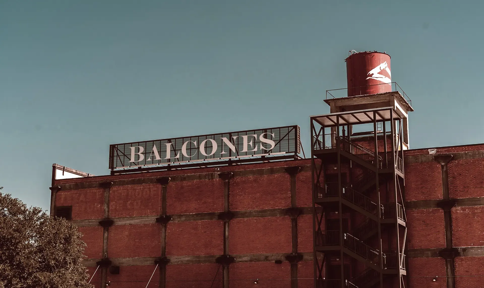 Red brick industrial building with a large BALCONES sign and an external metal staircase, next to a red water tower under a clear sky.