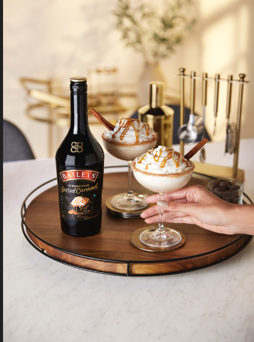 Botella Baileys sobre la mesa, dos copas con postre . todo sobre una bandeja de madera en una mesa.