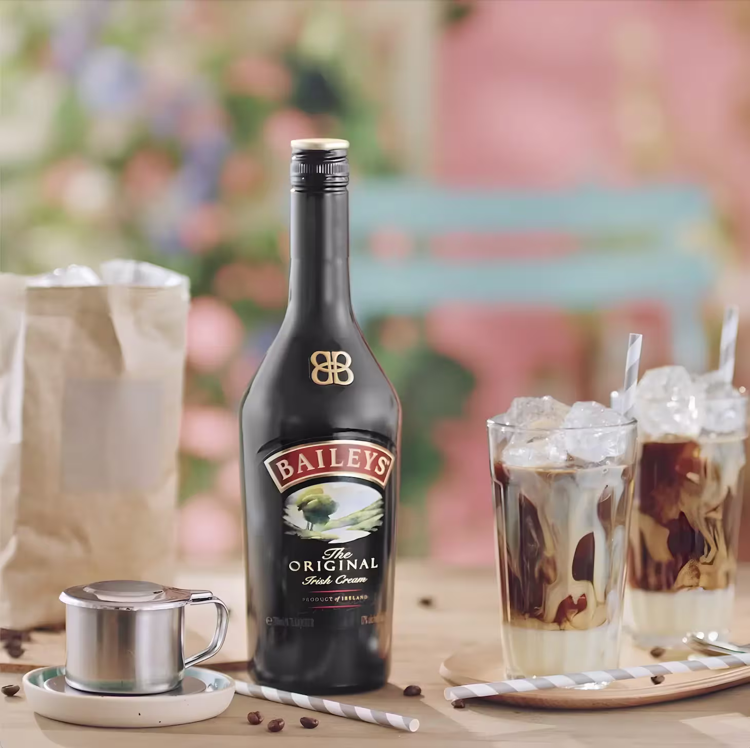 Una botella de Baileys Irish Cream sobre una mesa con dos bebidas de café helado, una taza de metal, una bolsa de papel y granos de café esparcidos.