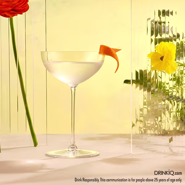 A cocktail in a coupe glass garnished with an orange twist sits on a light-colored surface, surrounded by yellow and red flowers and clear vertical panels.