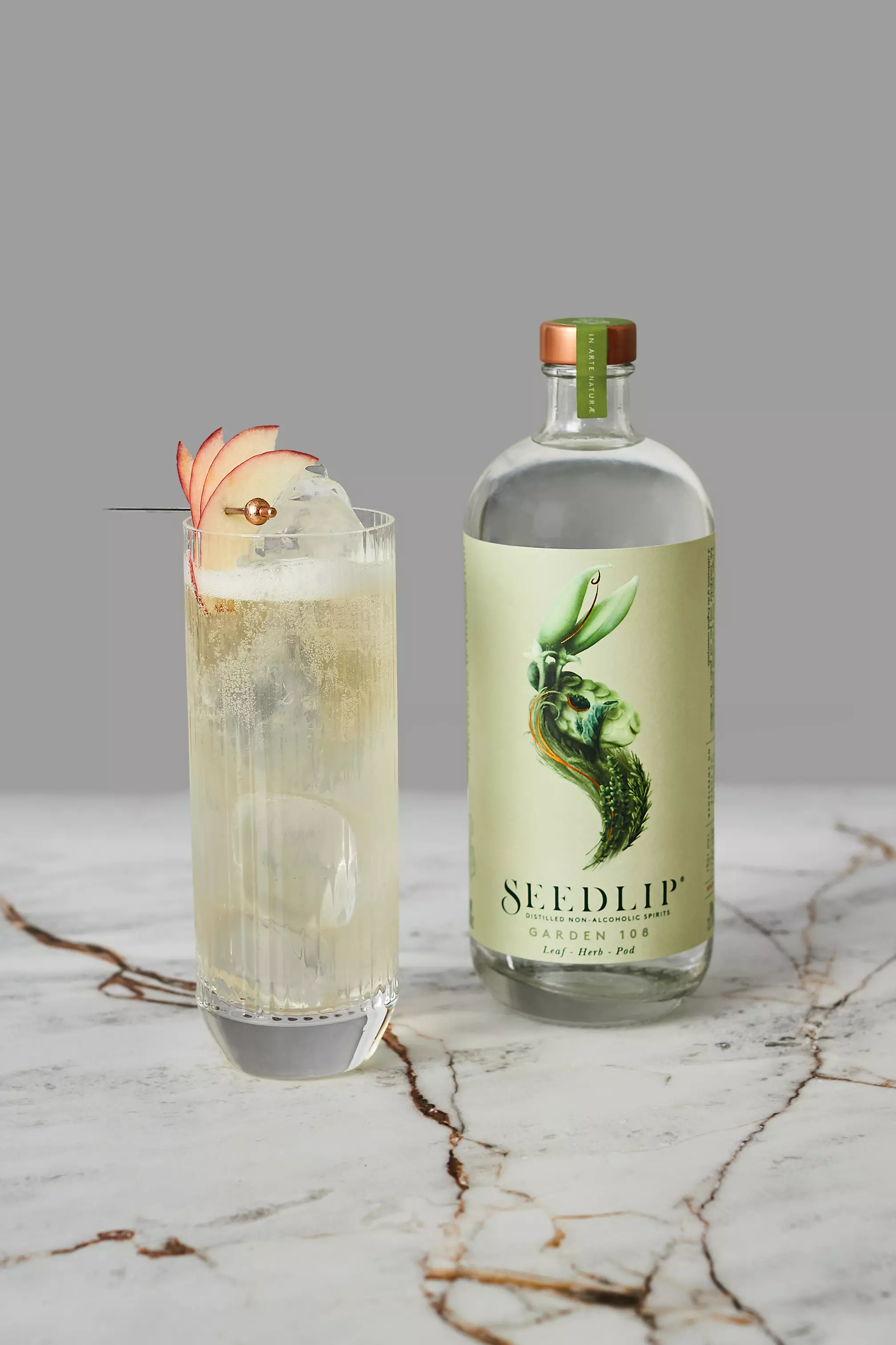 A glass of iced clear cocktail garnished with apple slices and a cherry sits next to a bottle of Seedlip Garden 108 non-alcoholic spirit on a marble surface.