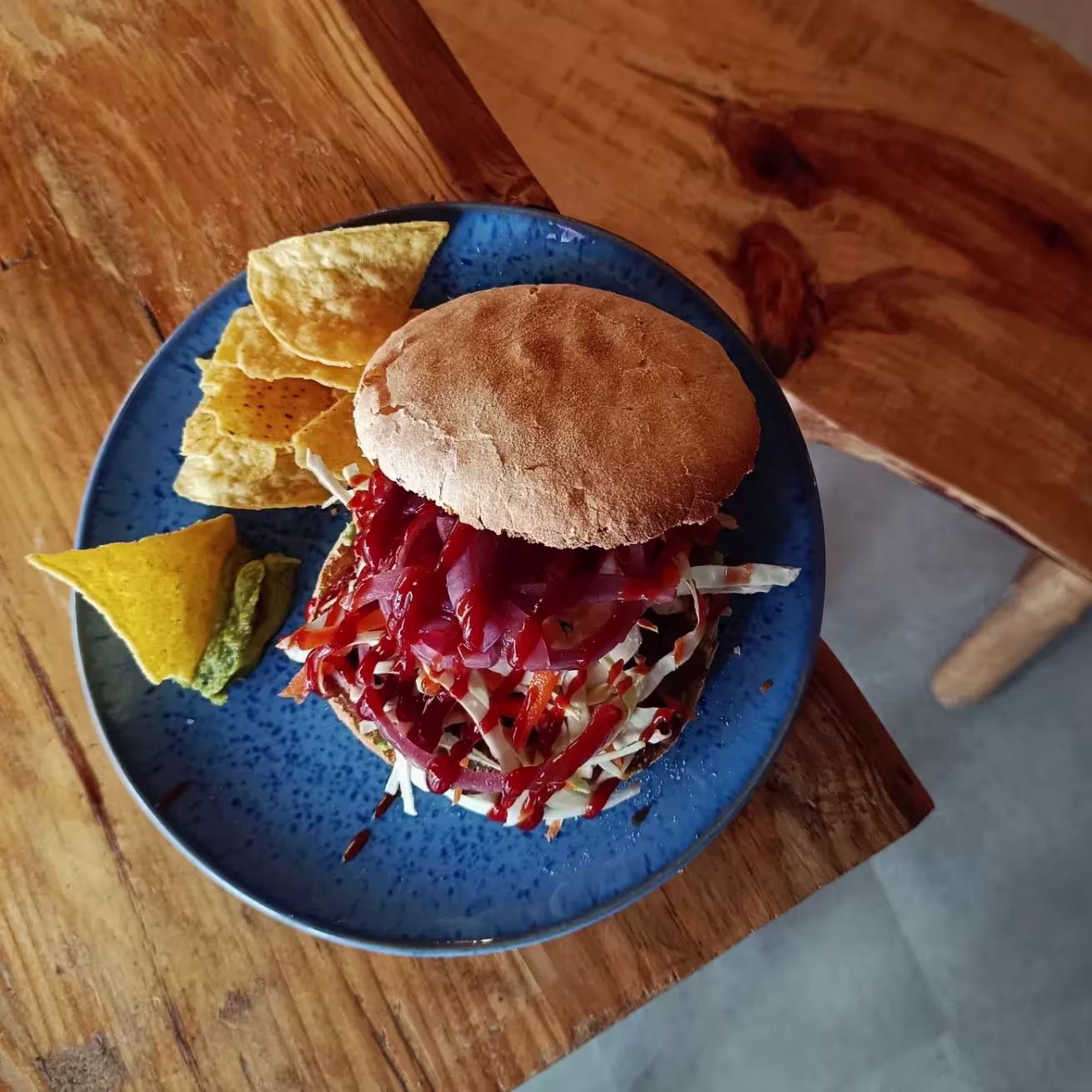 Hamburguesa con col lombarda, zanahoria rallada y salsa barbacoa en pan rústico, servida con nachos y guacamole sobre plato azul.