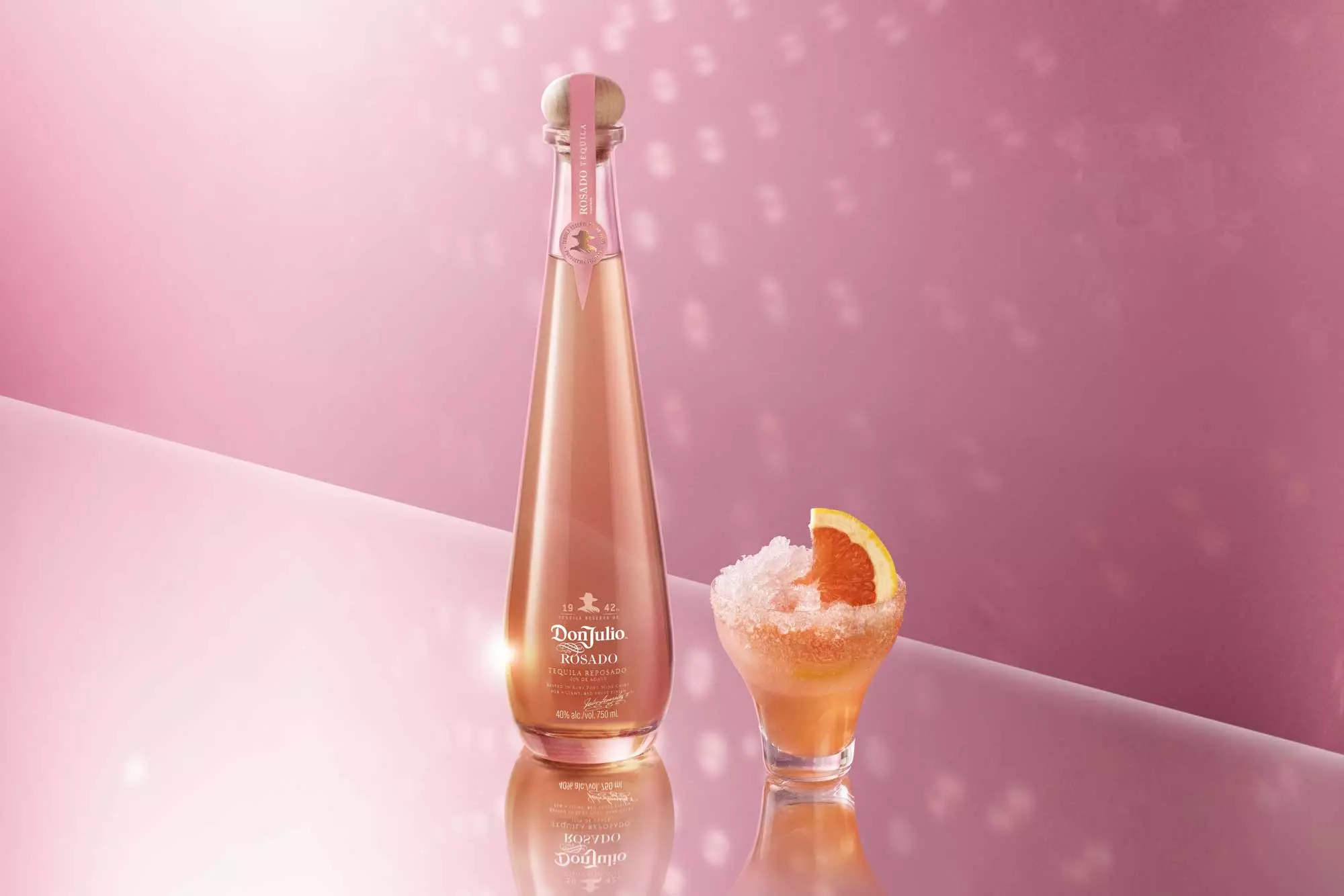A tall, elegant bottle of pink Don Julio Rosado tequila stands next to a short glass of pink cocktail with crushed ice and a grapefruit slice, set against a pink background.