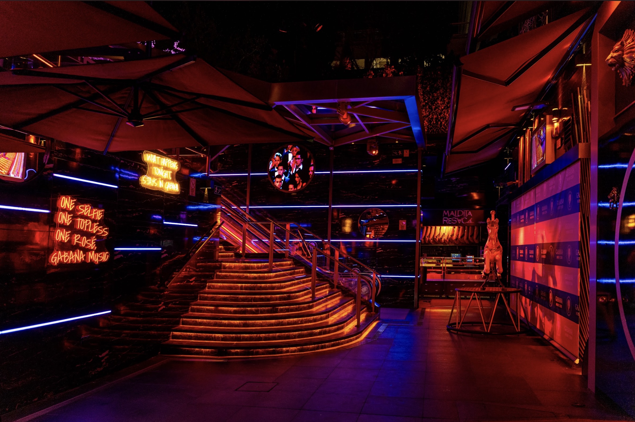 La entrada de Gabana destaca por su ambiente vibrante y lujoso, con luces de neón en tonos rojos y azules. Escaleras iluminadas y frases atrevidas en neón crean un entorno exclusivo y sofisticado que invita a una experiencia nocturna memorable.