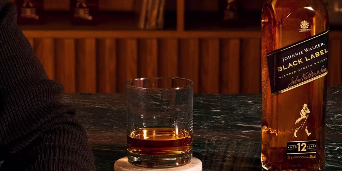 A man with a beard, wearing a dark blazer, holds a drink and looks at the camera in a warmly lit room with decorative letters on the wall behind him.