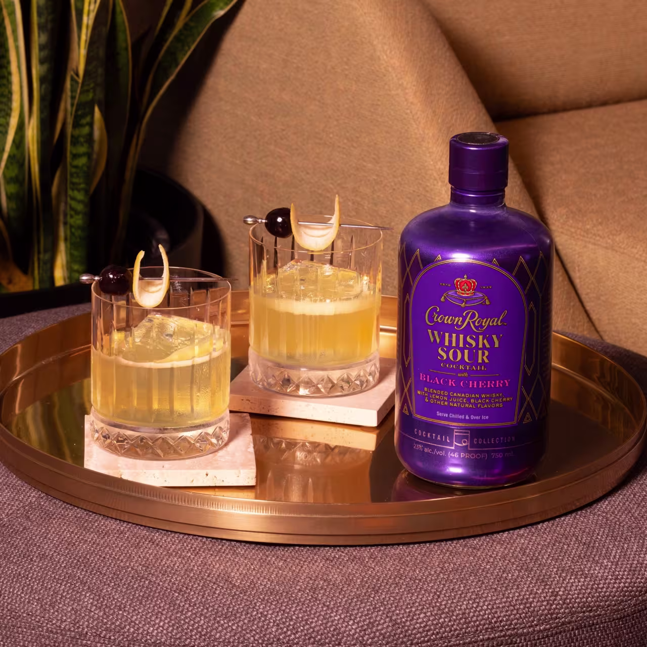 A gold tray holds two whisky sour cocktails with garnish and a purple bottle of Crown Royal Whisky Sour Black Cherry on a brown surface next to a green plant.