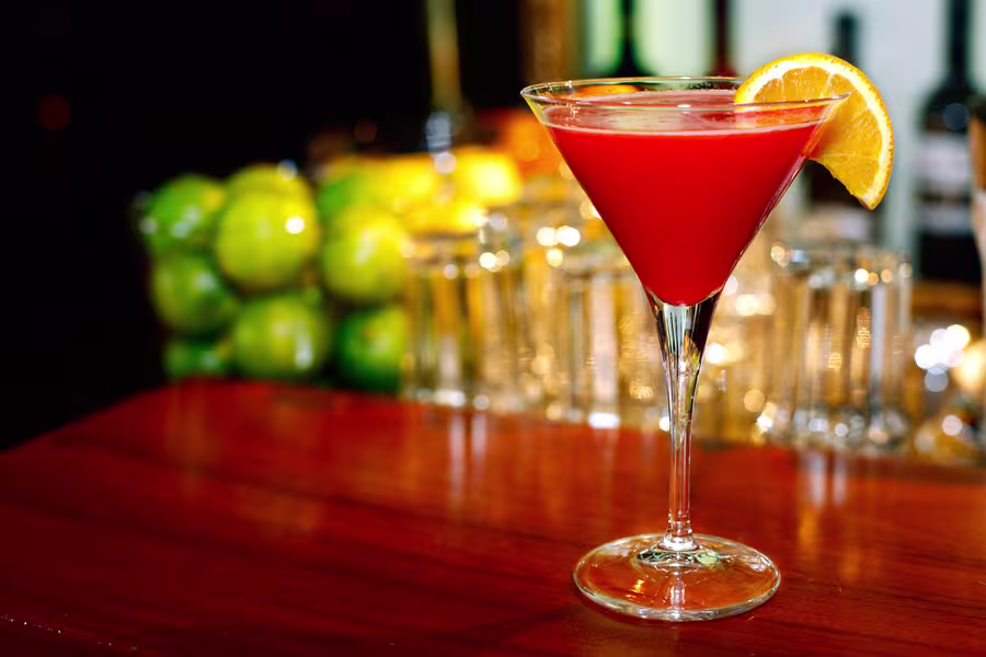 A red cocktail in a martini glass garnished with an orange slice sits on a wooden bar, with green apples and glassware in the background.
