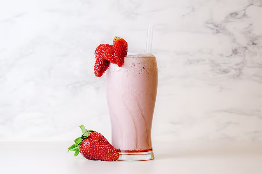 A glass of strawberry smoothie with a straw, garnished with two strawberries on the rim, and another strawberry resting at the base.