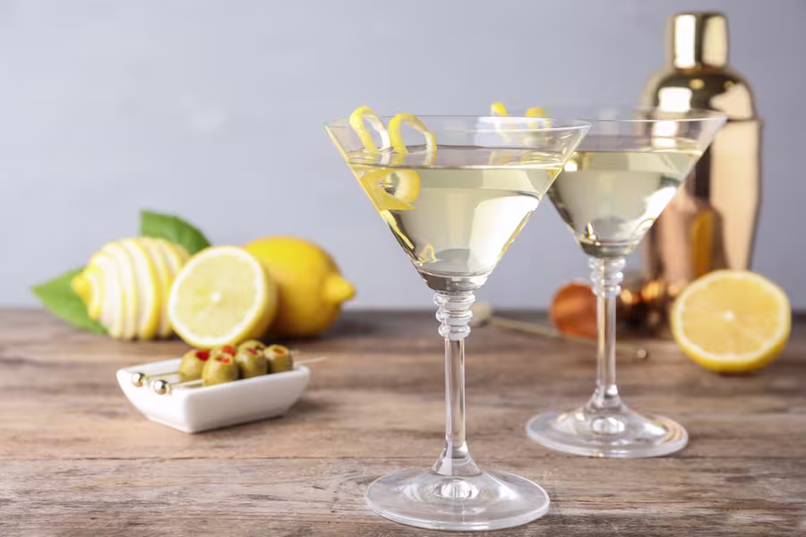 Two martini glasses with clear cocktails and lemon twists sit on a wooden surface, with lemons, olives, and a cocktail shaker in the background.