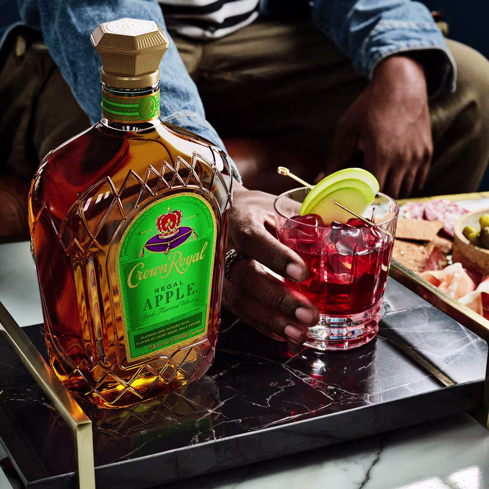 A bottle of Crown Royal Regal Apple whiskey next to a cocktail with ice and apple slices, being held by a person on a black tray with snacks in the background.