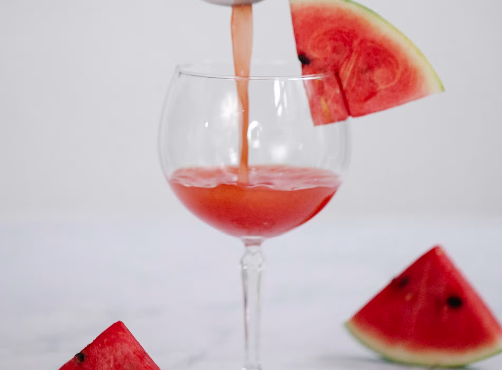 A glass is being filled with pink watermelon juice, garnished with a watermelon slice, and surrounded by triangular pieces of watermelon on a light surface.