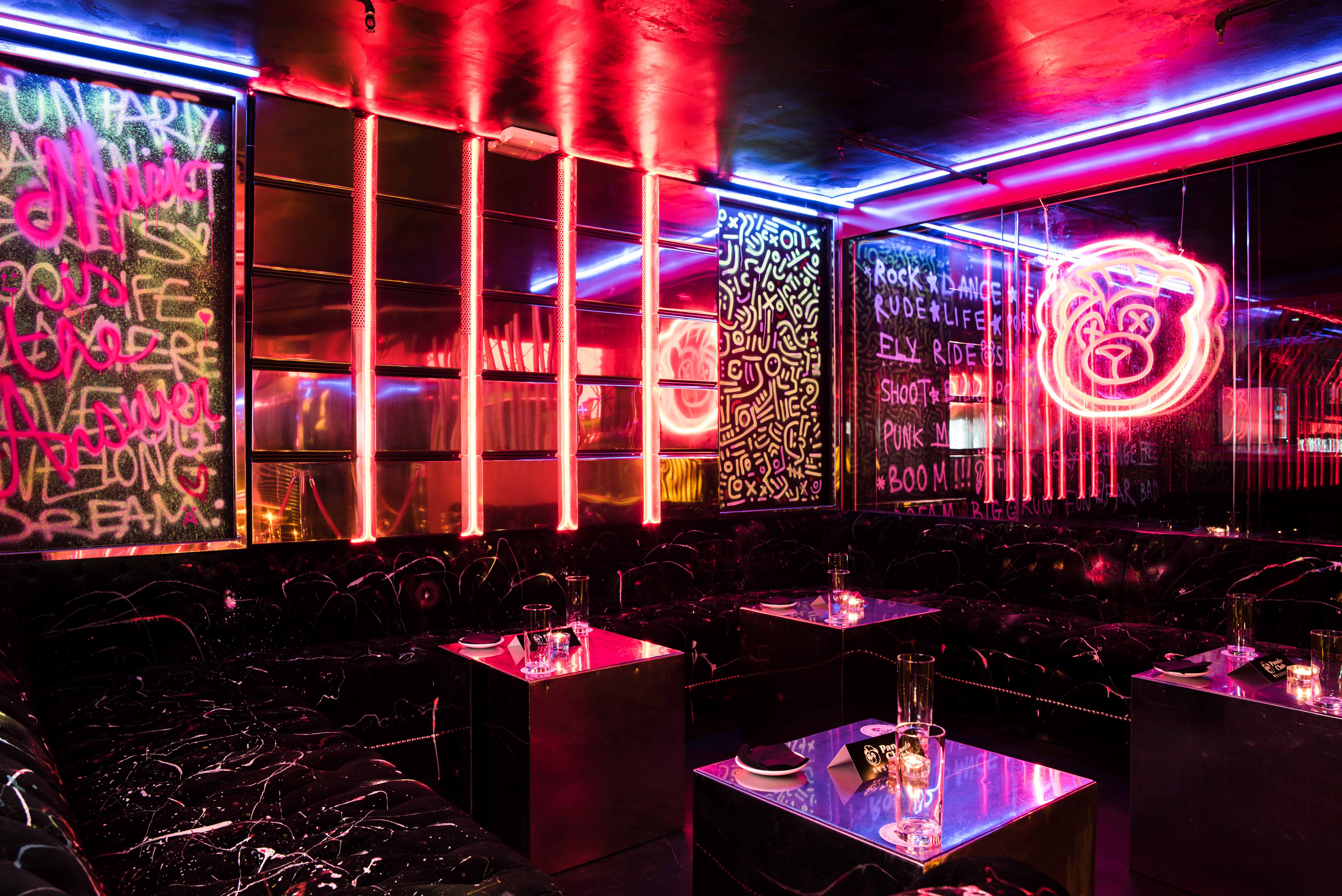 La imagen muestra el interior del Panda Club, con una decoración vibrante y moderna, llena de luces de neón en tonos rosas y azules. Graffitis y arte urbano adornan las paredes, creando un ambiente juvenil y energético, ideal para una noche de fiesta.