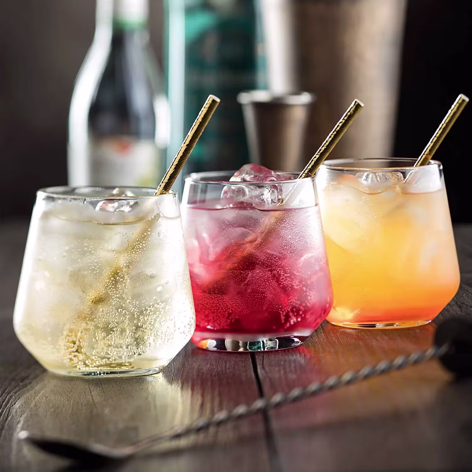 Three cocktails in short glasses with ice and metal straws sit on a wooden surface, each a different color: clear, red, and orange. A blurred bottle and bar tools are in the background.
