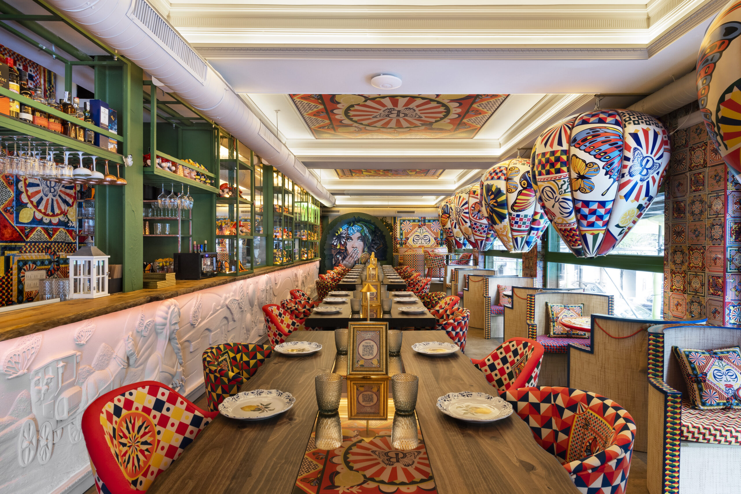 La imagen muestra el interior del restaurante Boom Boom Ciao. El techo presenta patrones intrincados con una mezcla de colores cálidos. La pared del fondo está llena de estantes con botellas. Cada mesa está adornada con patrones únicos y coloridos.