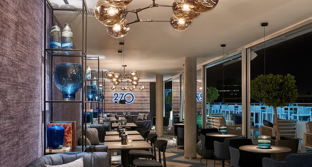 Interior elegante de 270º SkyBar en Hotel Barceló, con sofás grises, lámparas modernas, decoración en azul y vistas nocturnas desde su terraza acristalada.