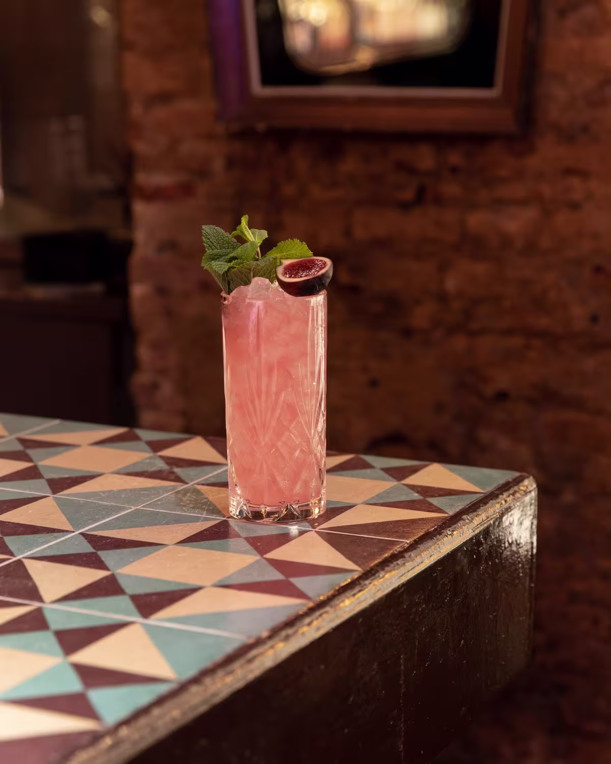 En Collage, un vibrante cóctel rosa servido en vaso tallado, adornado con menta fresca y una higo, refleja un toque fresco y artístico.