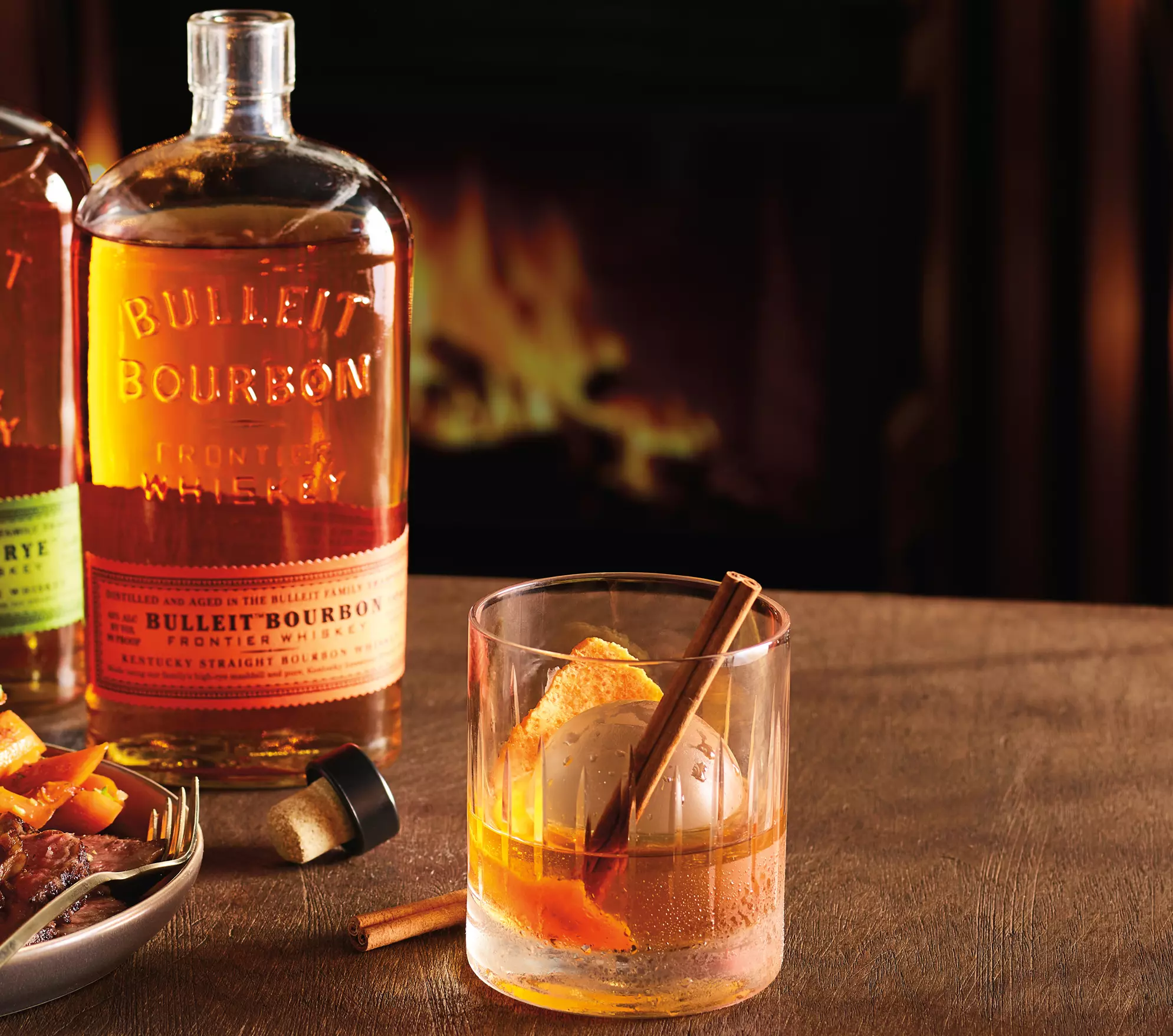A bottle of Bulleit Bourbon and a cocktail with ice, orange peel, and a cinnamon stick sit on a wooden table in front of a fireplace.