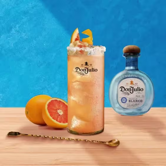 Cóctel Don Paloma junto a una botella de Don Julio Blanco, una toronja y media cortada y un batidor.