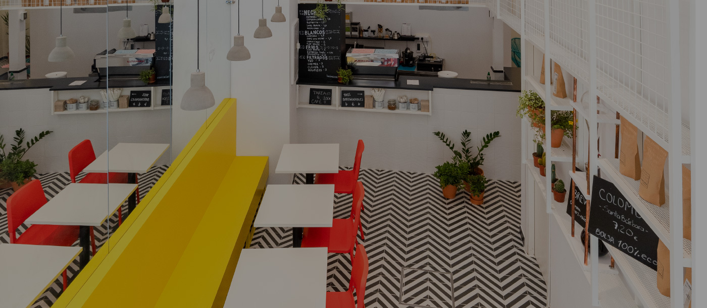 Interior de Parcería con suelo de zigzag blanco y negro, bancos amarillos, sillas rojas, plantas en macetas y barra con cartel de bebidas.