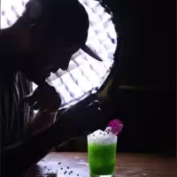 En la imagen se ve la sombra de una persona, mientras mira un cóctel de color verde manzana con detalles en la parte superior en blanco y morado.