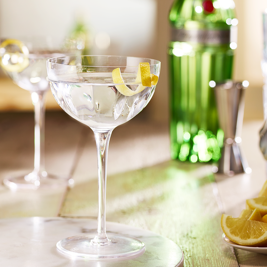 La imagen muestra dos copas rellenas con el cóctel Dry martini, de fondo, una botella de gin Tanqueray n° TEN y otros elementos de coctelería alrededor.