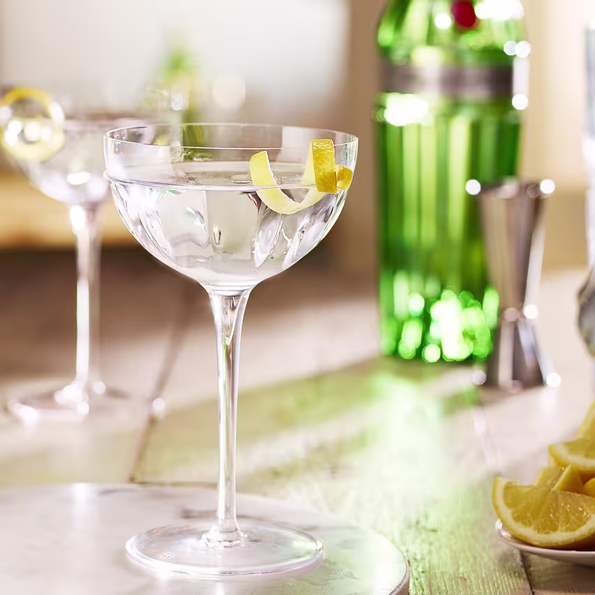 La imagen muestra dos copas rellenas con el cóctel Dry martini, de fondo, una botella de gin Tanqueray n° TEN y otros elementos de coctelería alrededor.