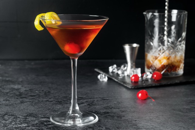 A martini glass with a dark cocktail, garnished with a lemon twist and a cherry, sits on a dark surface with a mixing glass, jigger, ice, and cherries in the background.