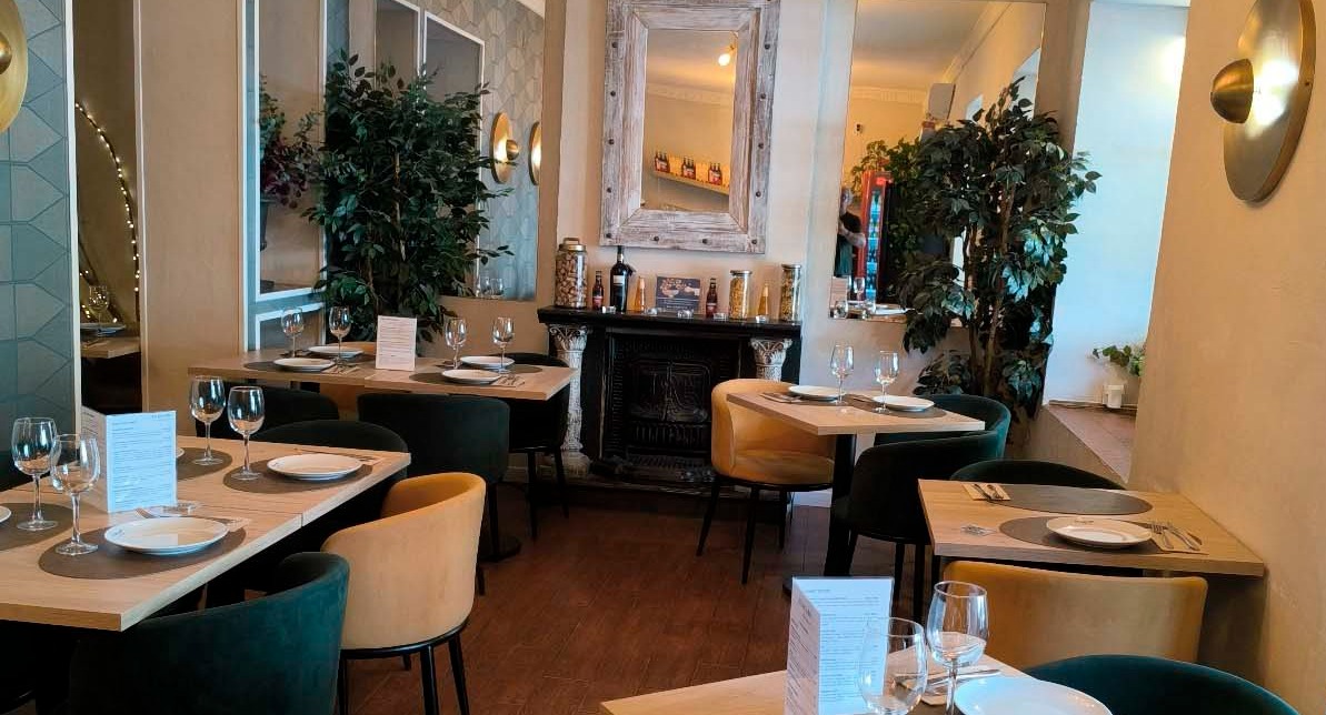 Salón acogedor con mesas vestidas, copas de vino, sillas de terciopelo y decoración con plantas y espejo en Bacaro.