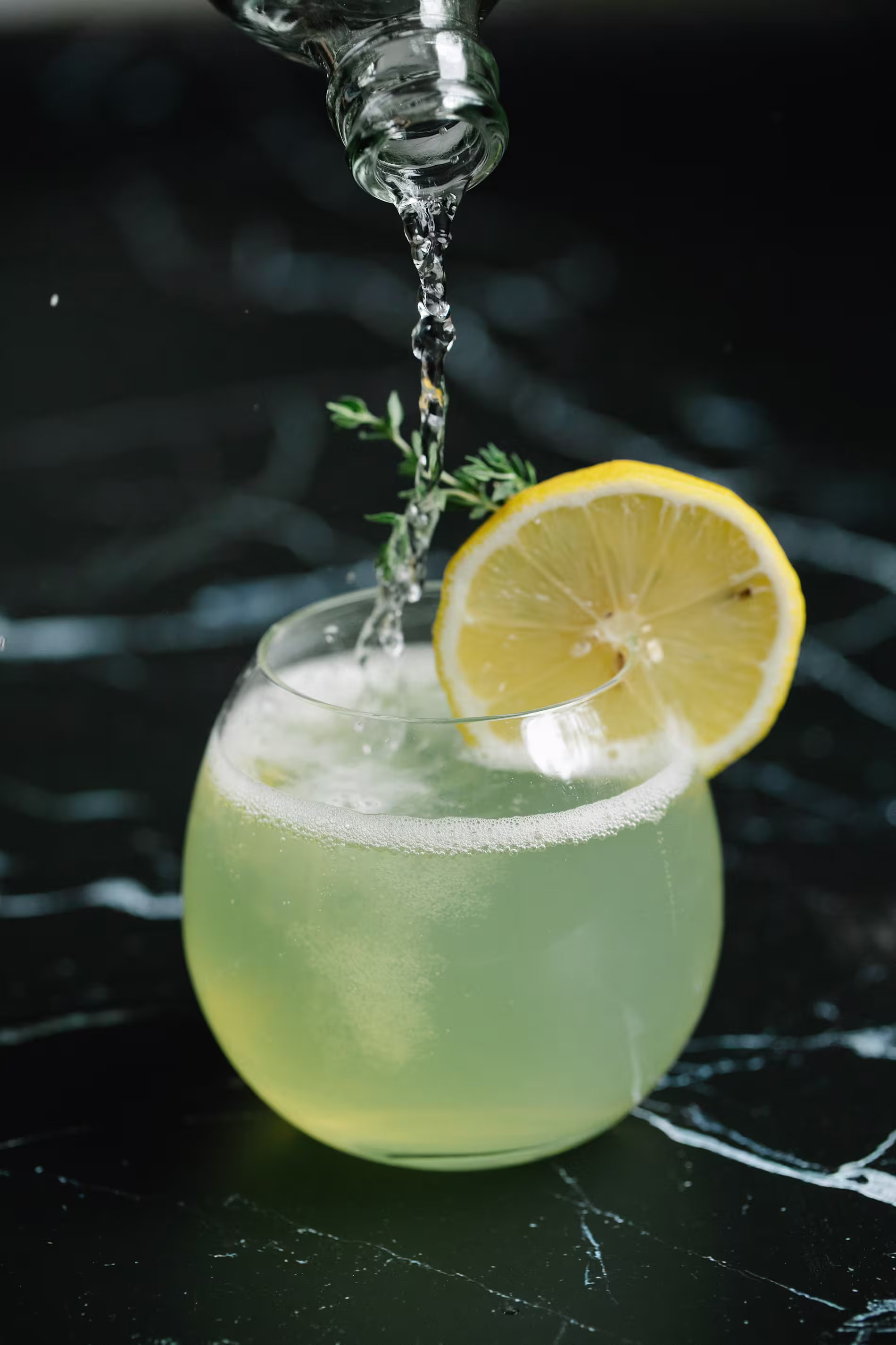 A clear liquid is being poured into a round glass of light yellow drink with a lemon slice and herb garnish on a dark marble surface.