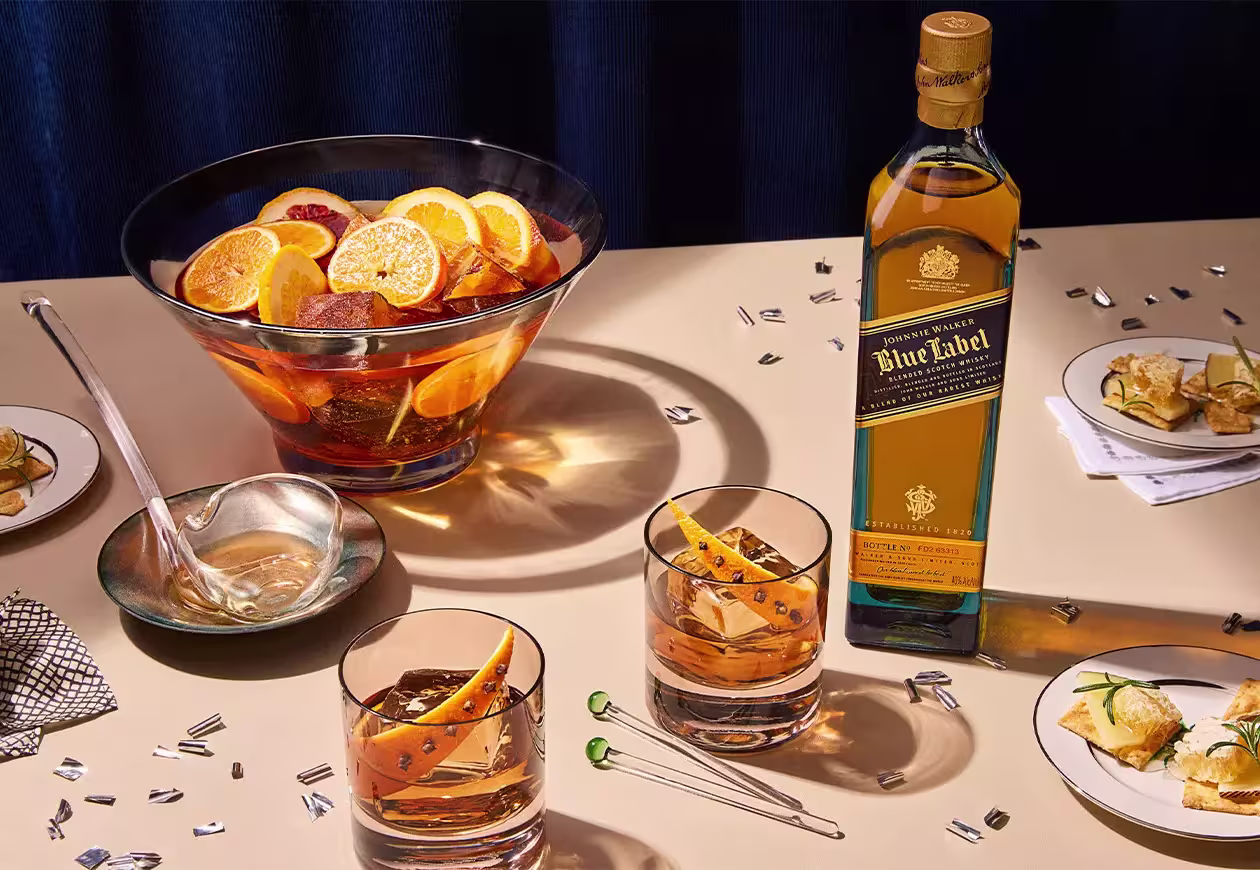 A bottle of Johnnie Walker Blue Label whisky sits on a table with two whisky glasses, a punch bowl with orange slices, and plates of appetizers.