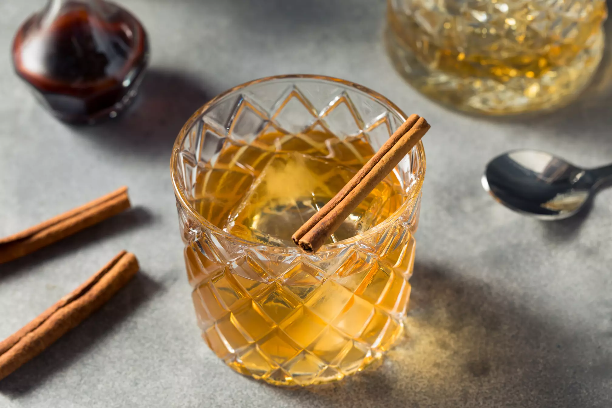 A glass of amber-colored cocktail with ice is garnished with a cinnamon stick, placed on a gray surface beside a spoon and extra cinnamon sticks.