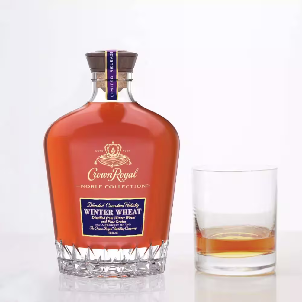 A bottle of Crown Royal Noble Collection Winter Wheat whisky stands next to a glass containing a small pour of whisky on a white background.
