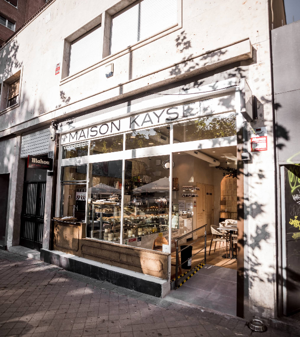 Fachada acristalada de Maison Kayser Padre Damián con vista directa al interior y al expositor de bollería artesanal junto a mesas bajas.