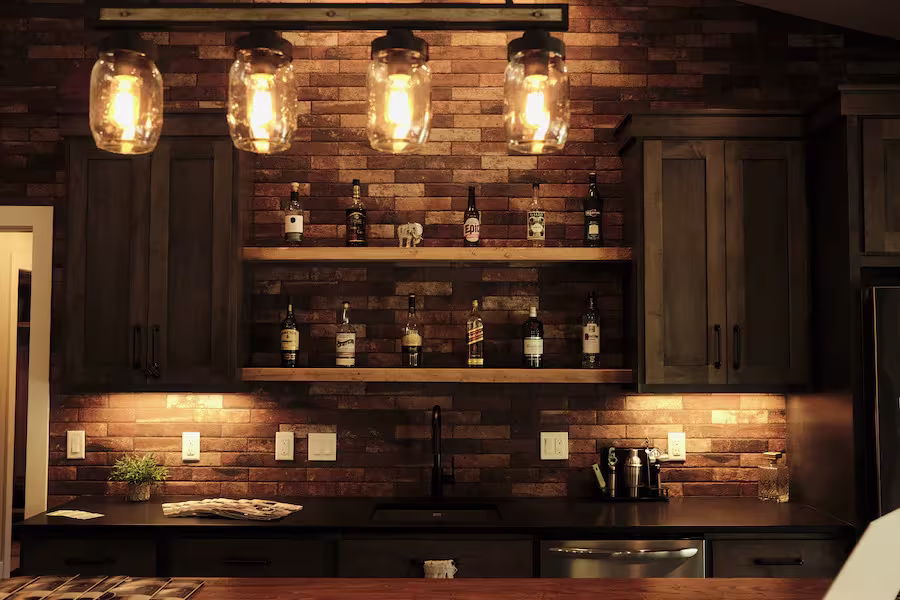Dimly lit kitchen with brick walls, wooden cabinets, shelves displaying bottles, and industrial-style hanging lights above a black countertop.