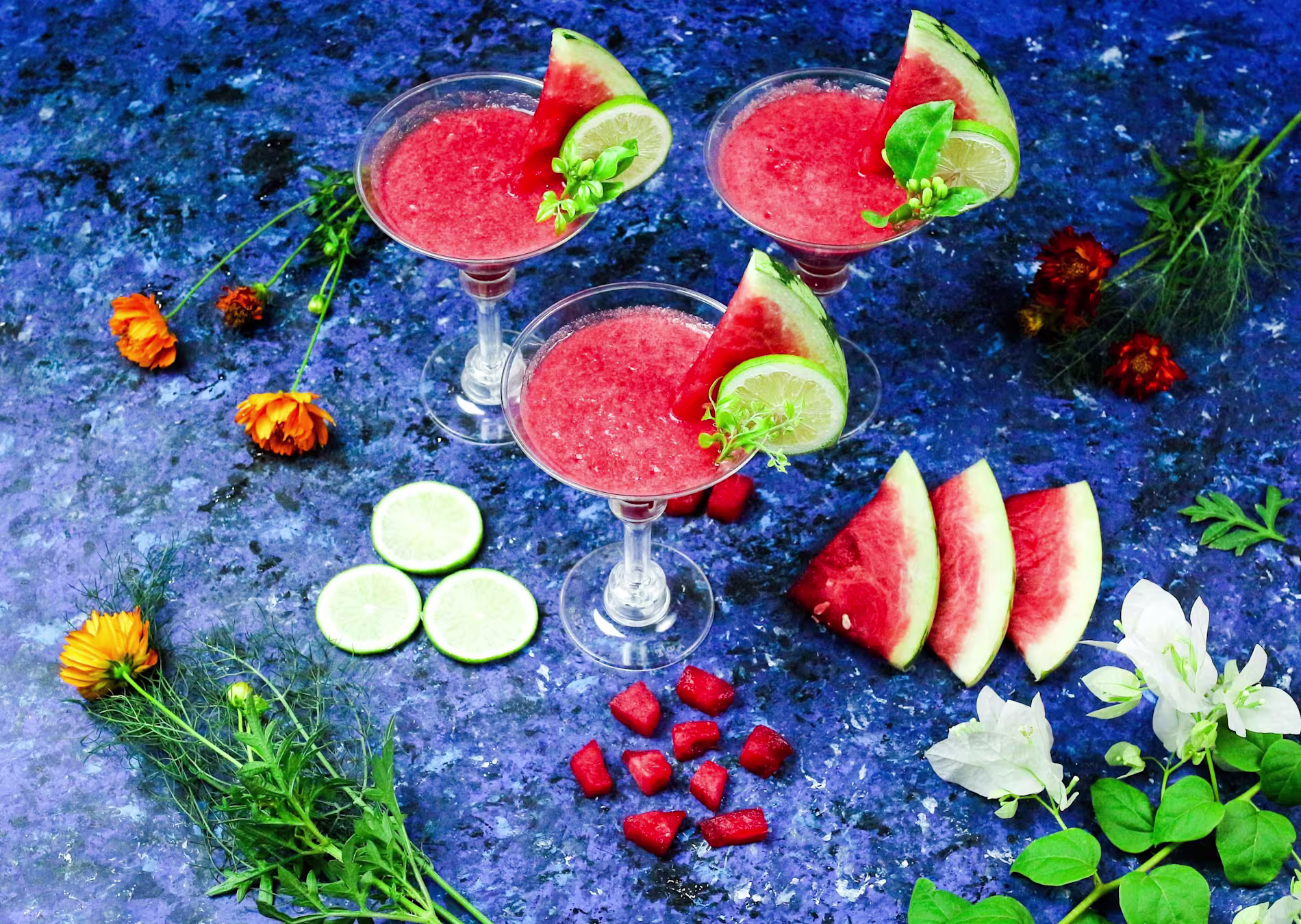 Three watermelon cocktails garnished with lime and mint on a blue surface, surrounded by watermelon slices, lime slices, flower blossoms, and green herbs.