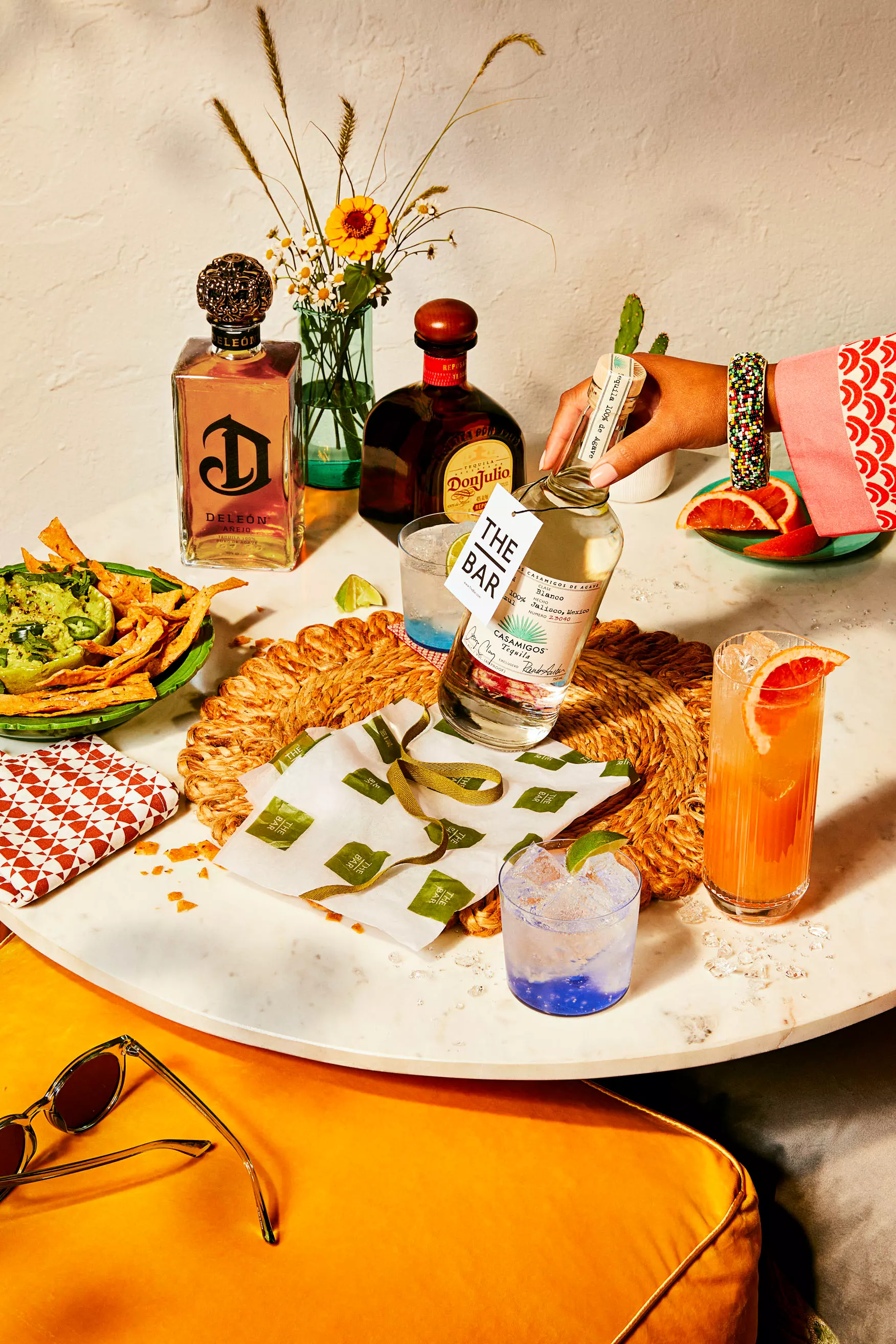 A round table with tequila bottles, cocktails, guacamole with chips, sunglasses, and a hand pouring liquor, set against a white wall with flowers in a vase.